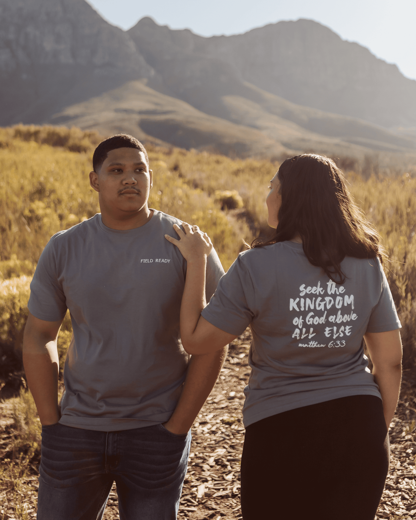 Two people standing in a field with mountains in the background, one wearing a t-shirt with text on the back.