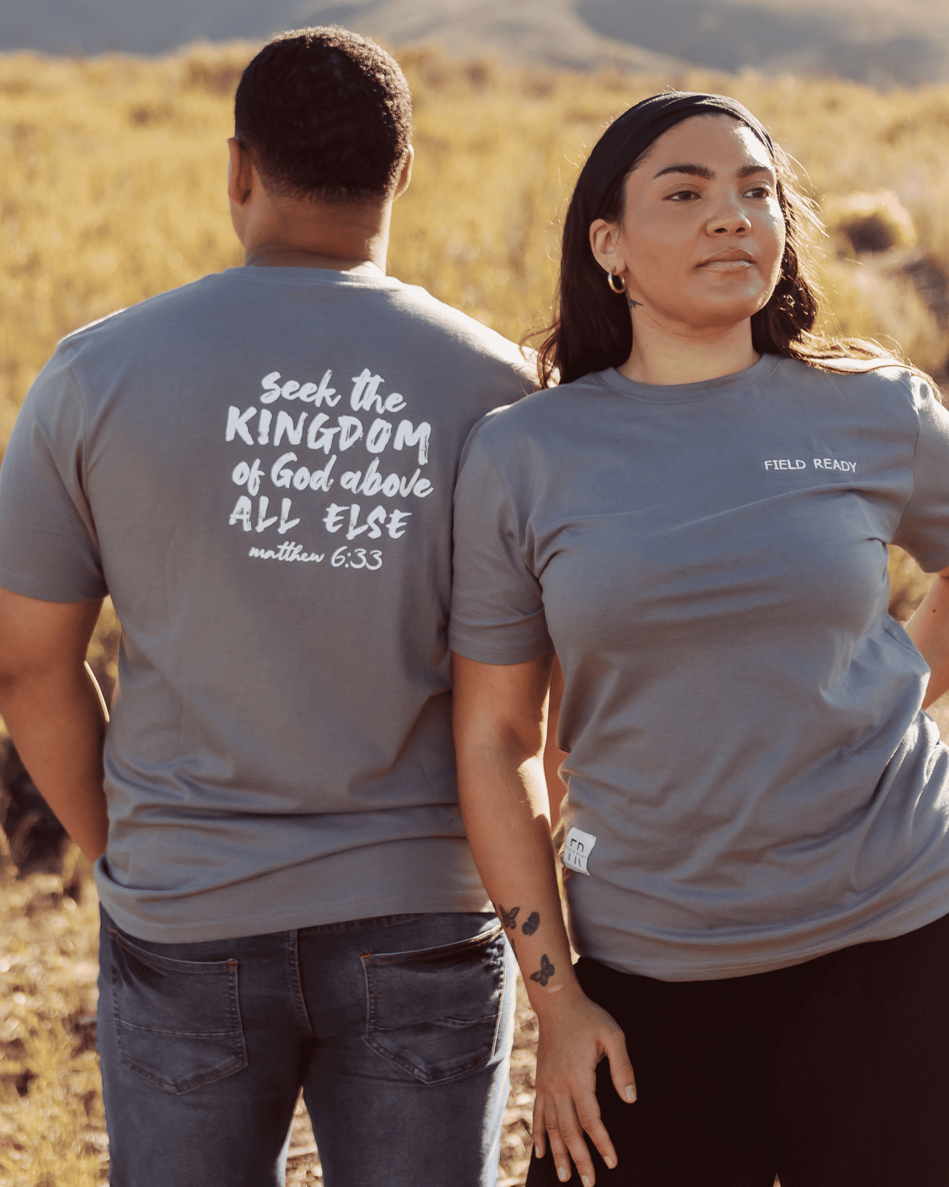 Two people wearing gray t-shirts with text, standing in a desert landscape.